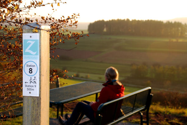 Bild vergrößern: Eine junge Frau sitzt an der Sitzgruppe oberhalb der Marbeckhänge. Im Vordergrund ist ein Markierungsposten mit einem blauen Schild der Station 8 des GeoPfads zu sehen.