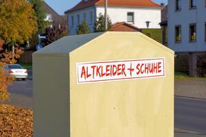 Bild vergrößern: Symbolbild Altkleidercontainer
Im Vordergrund des Bildes ist ein Altkleidercontainer mit einer roten Aufschrift "Altkleider + Schuhe" abgebildet. Der Container steht in einem Wohngebiet an einer Straße. Im Hintergrund sind Wohnhäuser und ein Auto zu sehen.