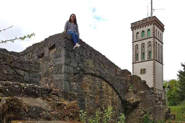 Bild vergrößern: Eine junge Frau sitzt auf den Mauern der Burgruine Eisenberg. Im Hintergrund ist der Georg-Viktor-Aussichtsturm zu sehen.